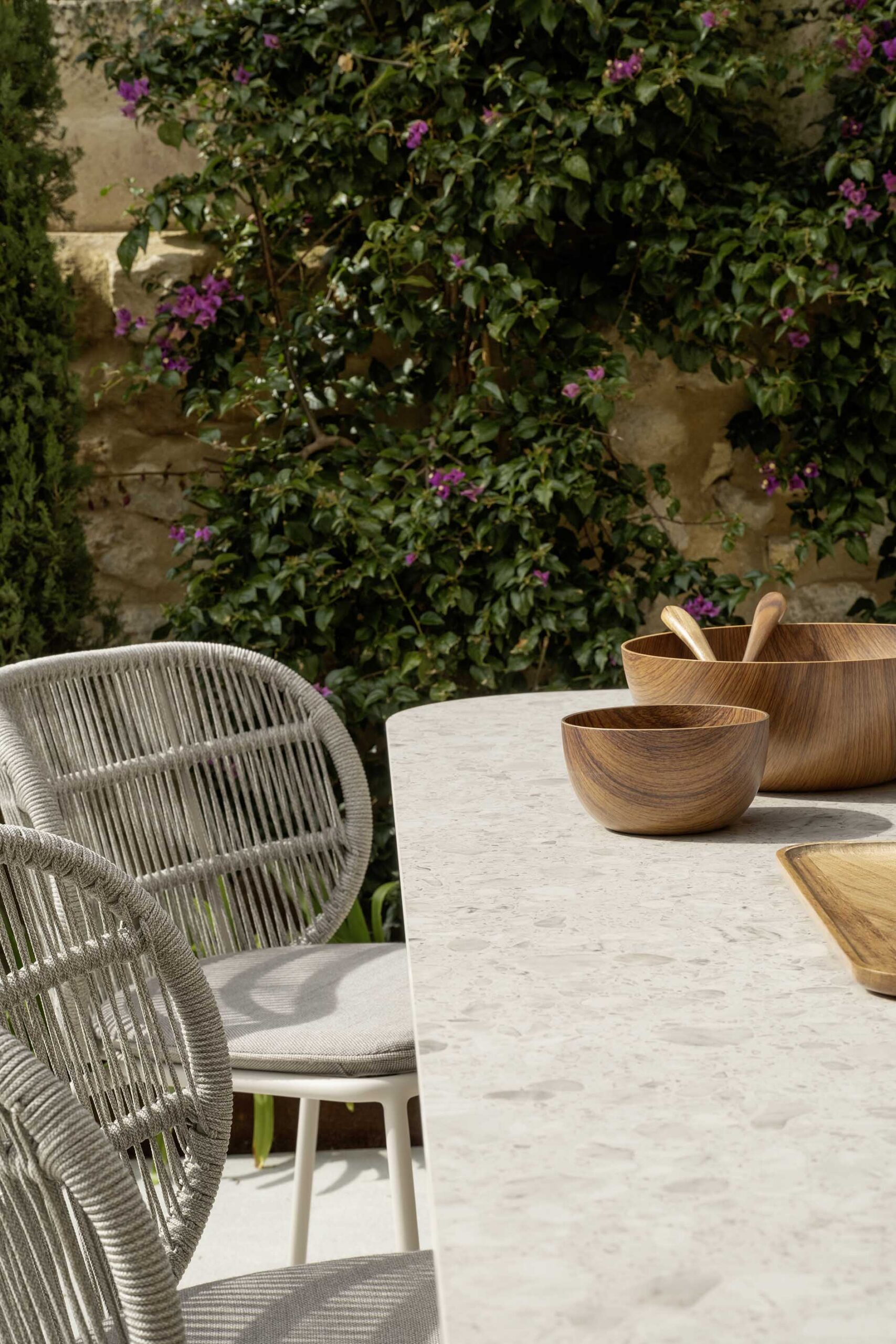 Detail of an outdoor table with light tabletop, wooden bowls, rope chairs and fuchsia flowers