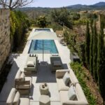 Aerial view of the terrace with pool, sun loungers, sofas, cypress trees and mountains in the background
