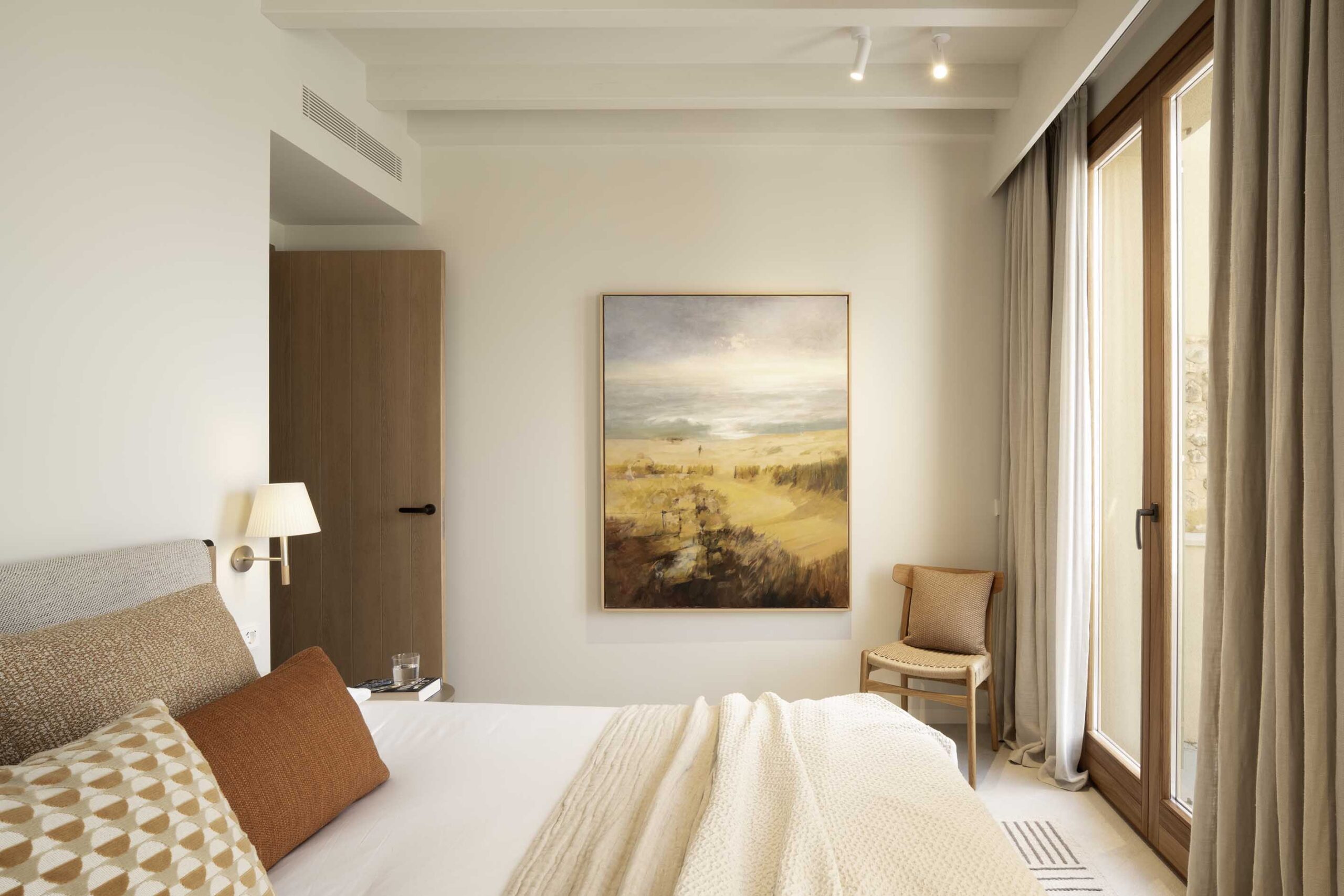 Bedroom with landscape painting, sand-coloured curtains, wooden door and large window with natural light