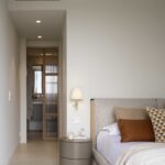 Bedroom with grey headboard, terracotta cushions, round bedside table and en suite bathroom in the background