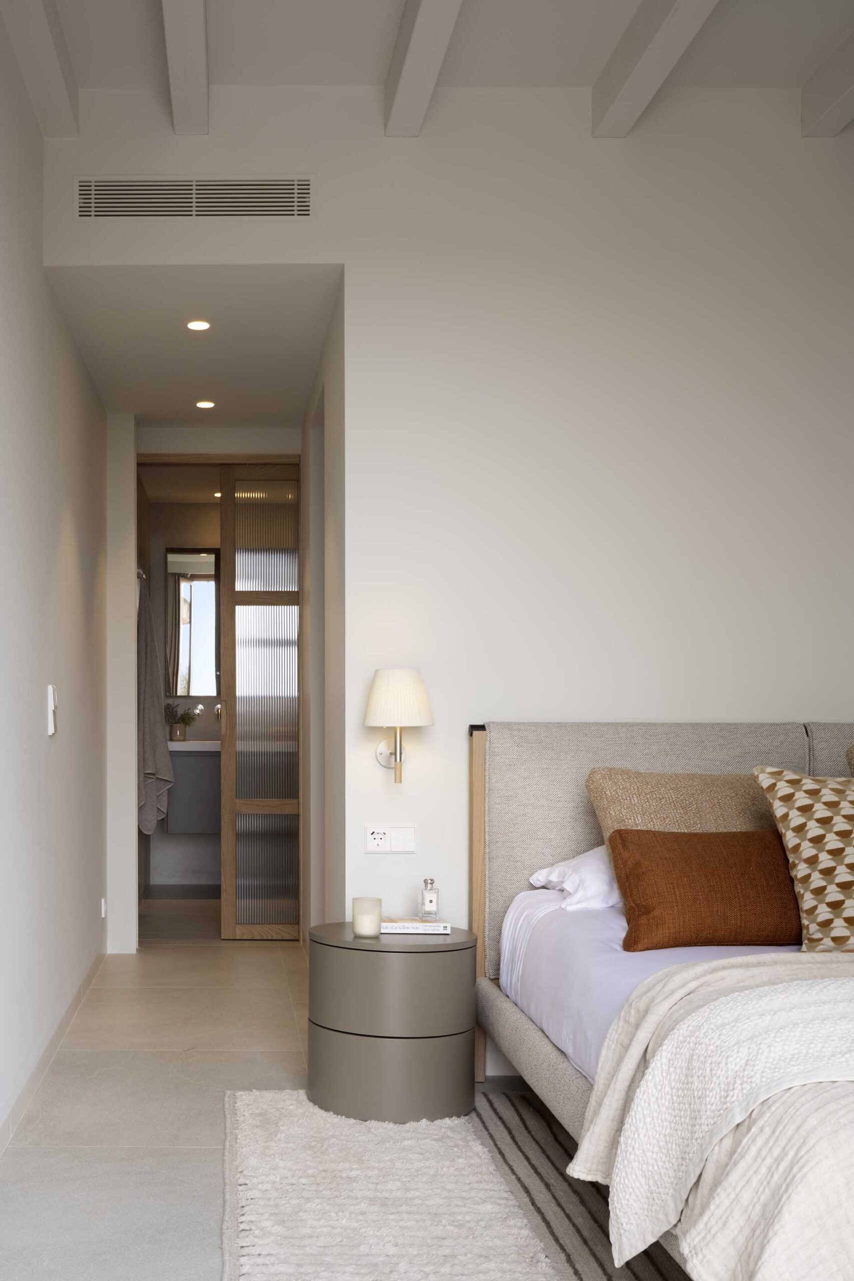 Bedroom with grey headboard, terracotta cushions, round bedside table and en suite bathroom in the background