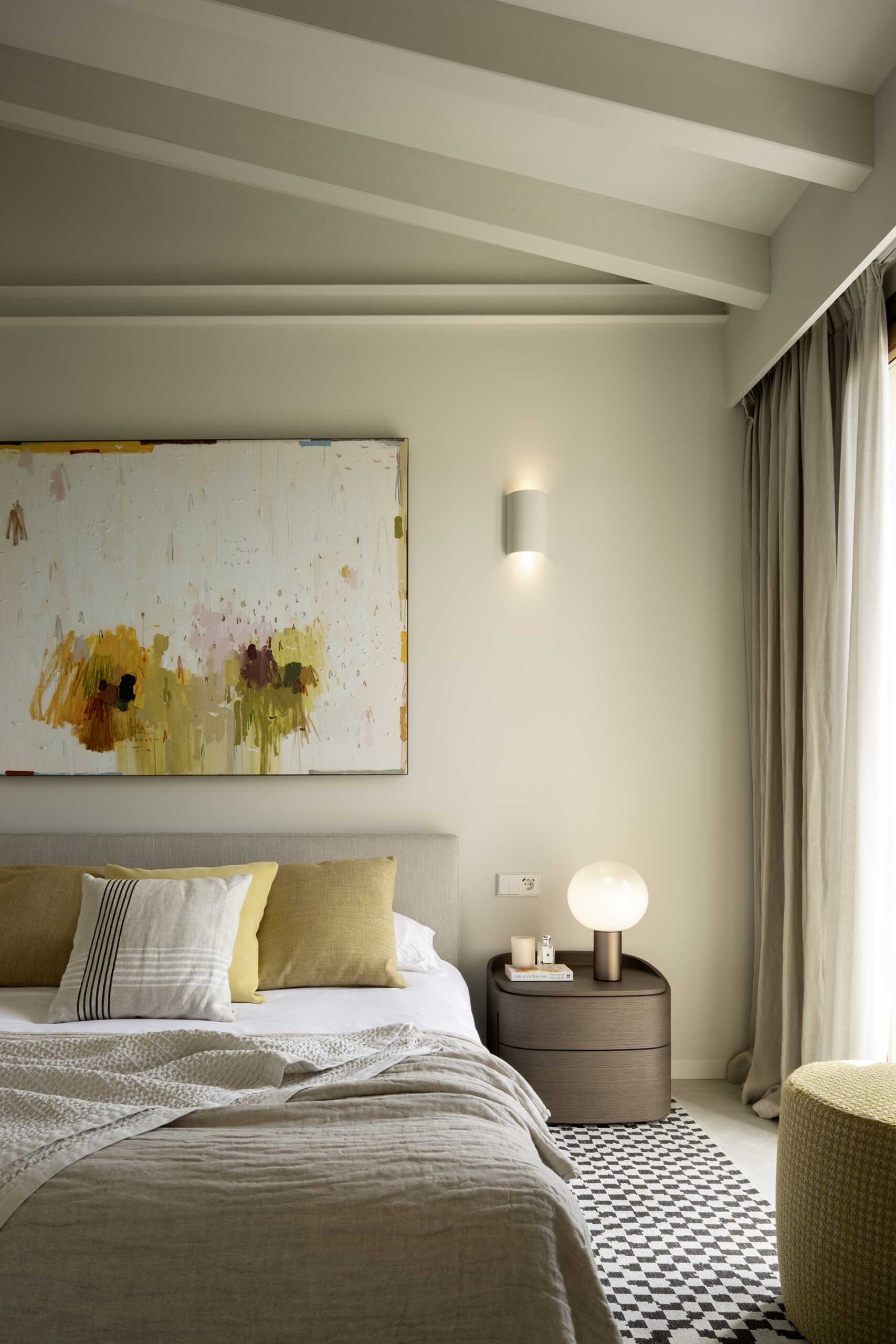 Bedroom with yellow-accented abstract painting, mustard cushions, bedside table with lamp and chequerboard rug