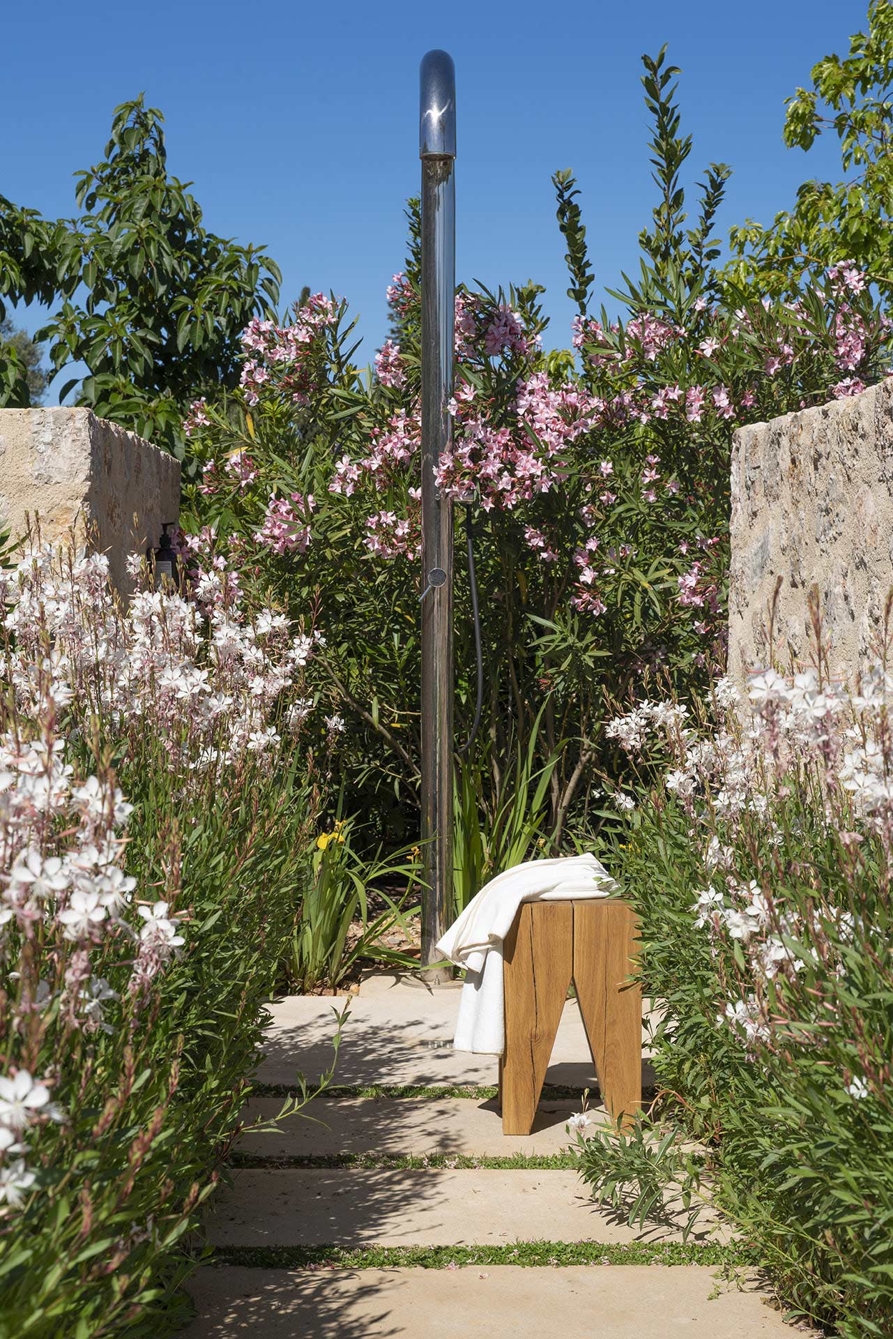 Detalle de la ducha exterior metálica con taburete de madera y toalla entre flores
