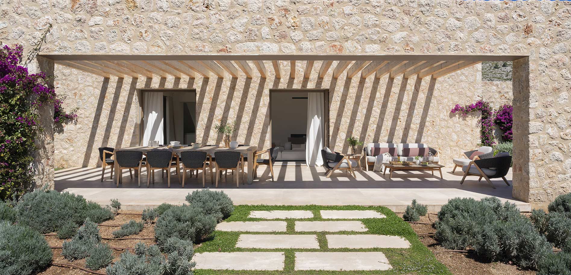 Porche con comedor y zona de estar bajo pérgola, con sombras sobre el muro de piedra