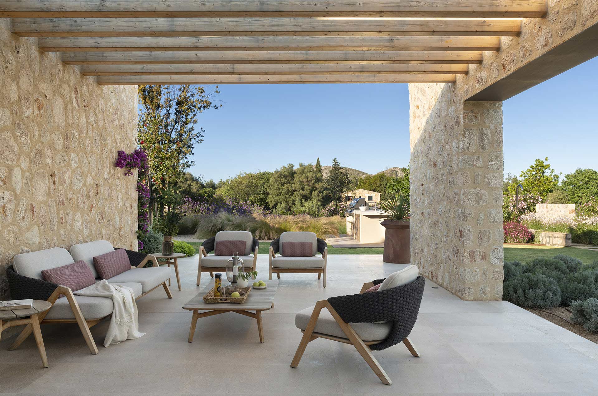 Zona de estar exterior con sofá y butacas de fibra bajo pérgola, entre muros de piedra