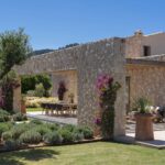 Esquina de la casa de piedra con olivo, buganvilla y porche con butacas junto al césped