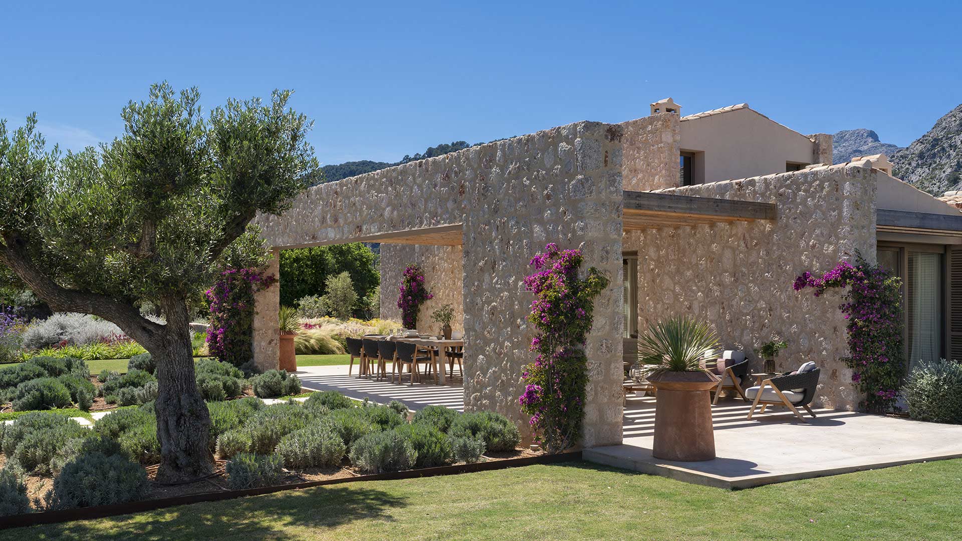 Esquina de la casa de piedra con olivo, buganvilla y porche con butacas junto al césped