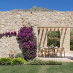 Muro de piedra con buganvilla y abertura que enmarca una mesa exterior, con la montaña detrás