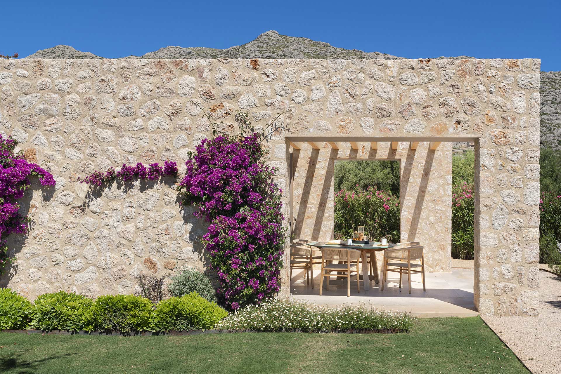 Muro de piedra con buganvilla y abertura que enmarca una mesa exterior, con la montaña detrás