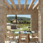 Mesa exterior con sillas de madera enmarcada por el hueco de piedra, con cocina exterior al fondo