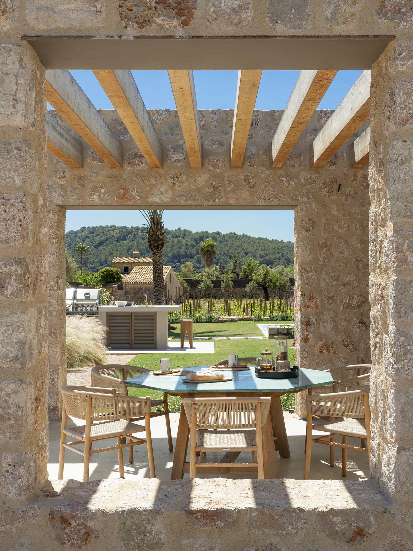 Mesa exterior con sillas de madera enmarcada por el hueco de piedra, con cocina exterior al fondo