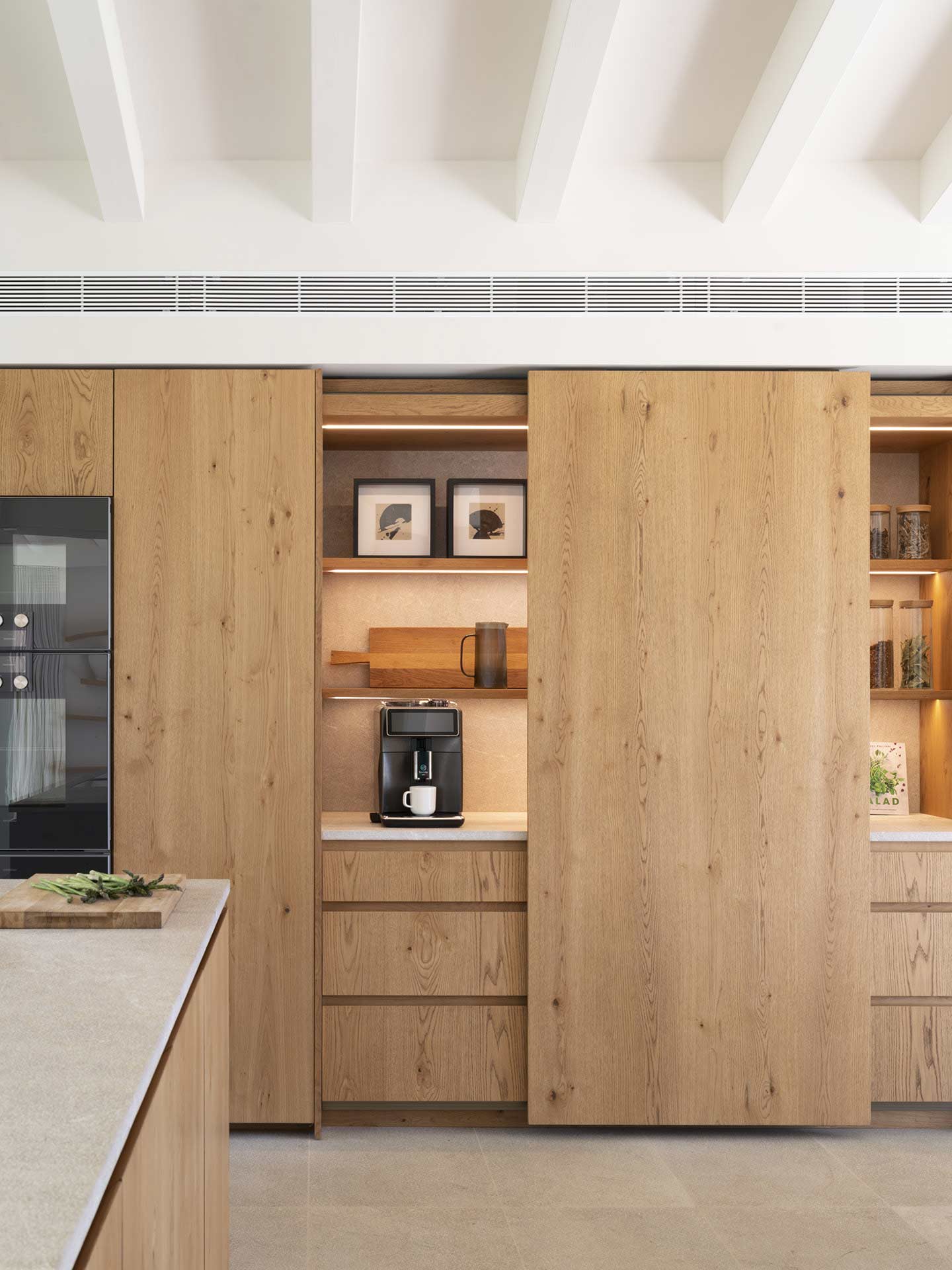 Armarios de madera con puerta corredera abierta que deja ver cafetera y estantes iluminados