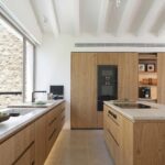 Cocina de madera con isla y encimera de piedra clara, hornos negros y gran ventanal