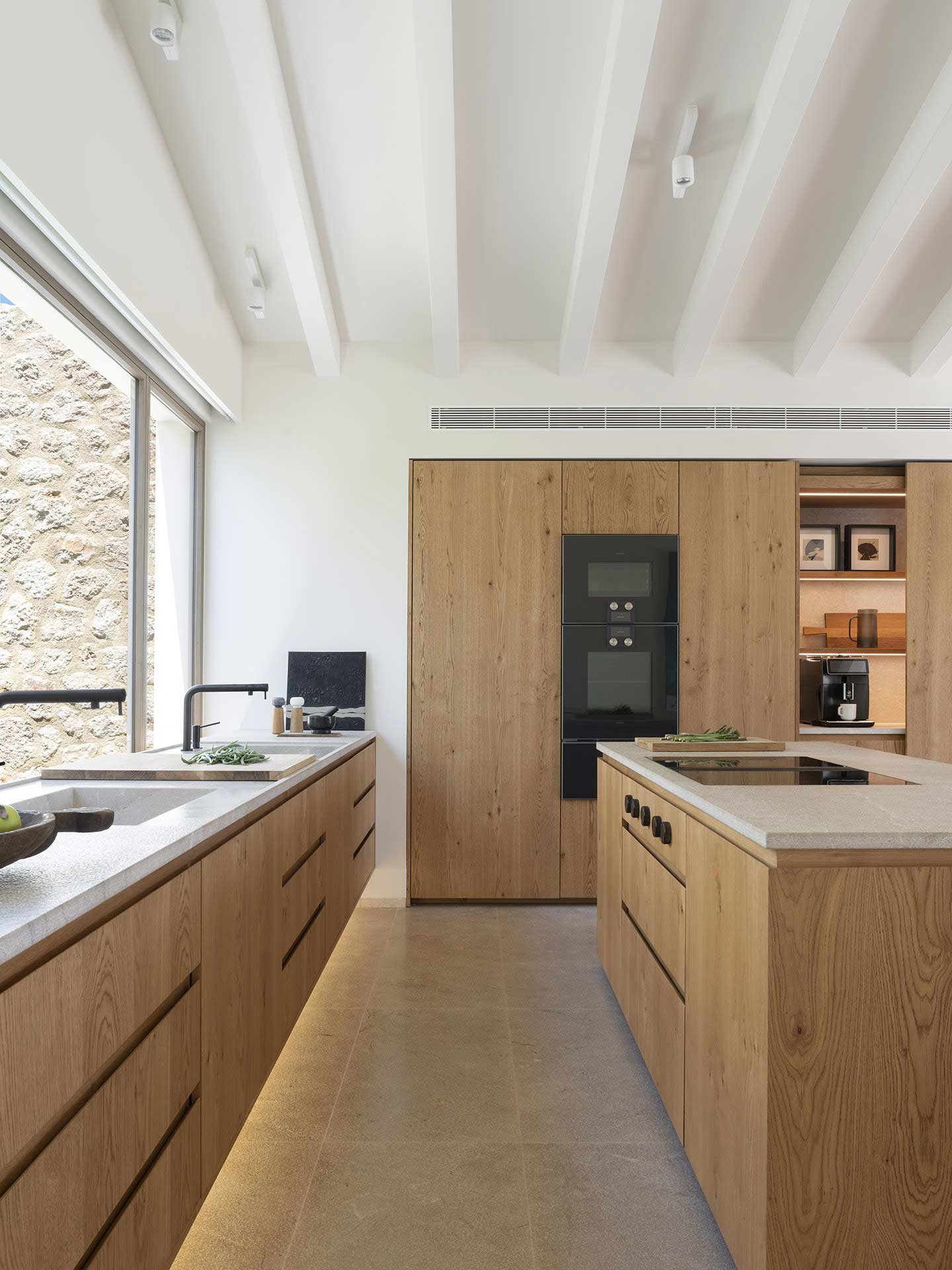Cocina de madera con isla y encimera de piedra clara, hornos negros y gran ventanal