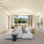Salón con techo de vigas blancas, sofá modular claro, chimenea y vistas a la montaña
