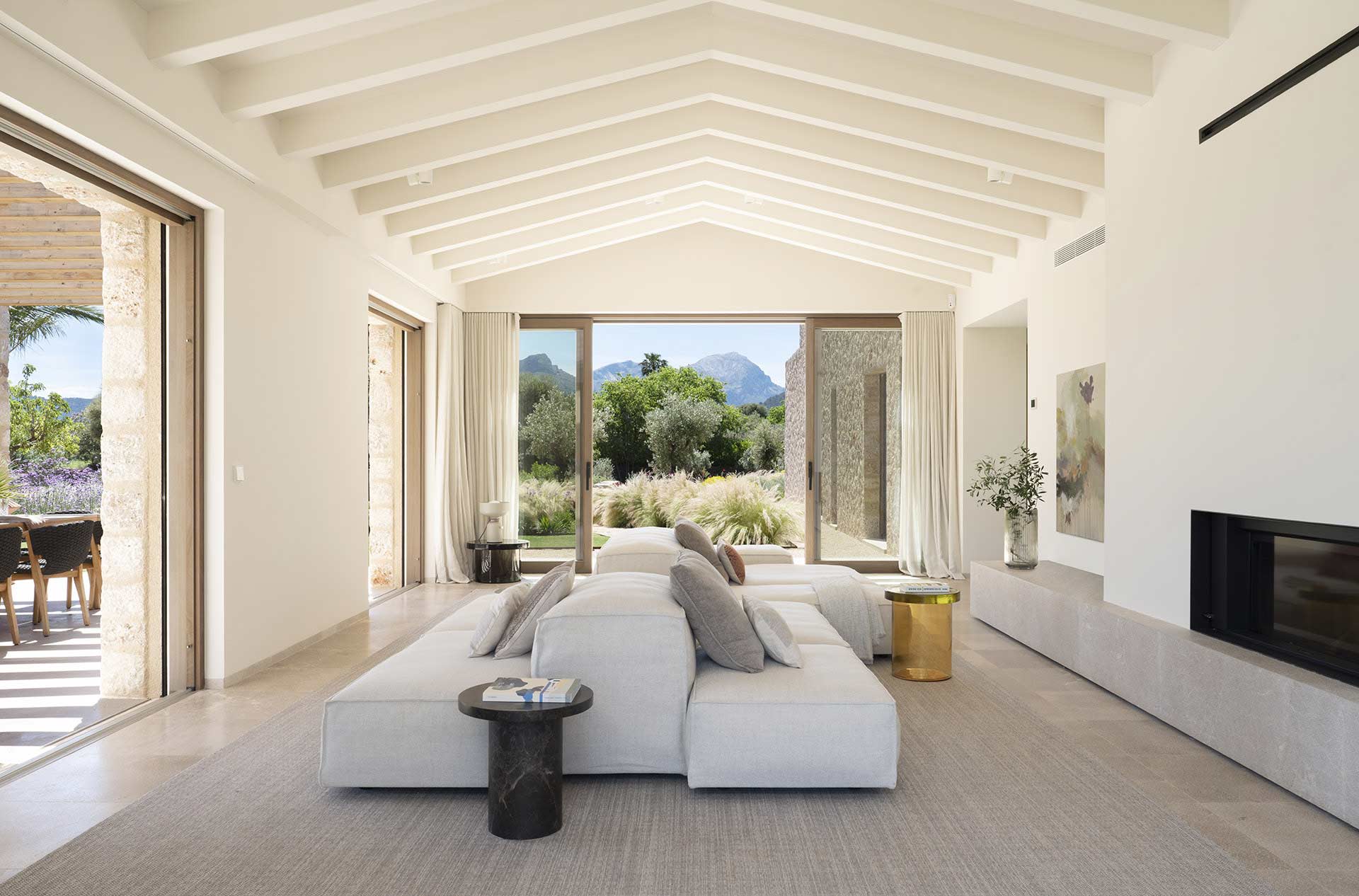 Salón con techo de vigas blancas, sofá modular claro, chimenea y vistas a la montaña