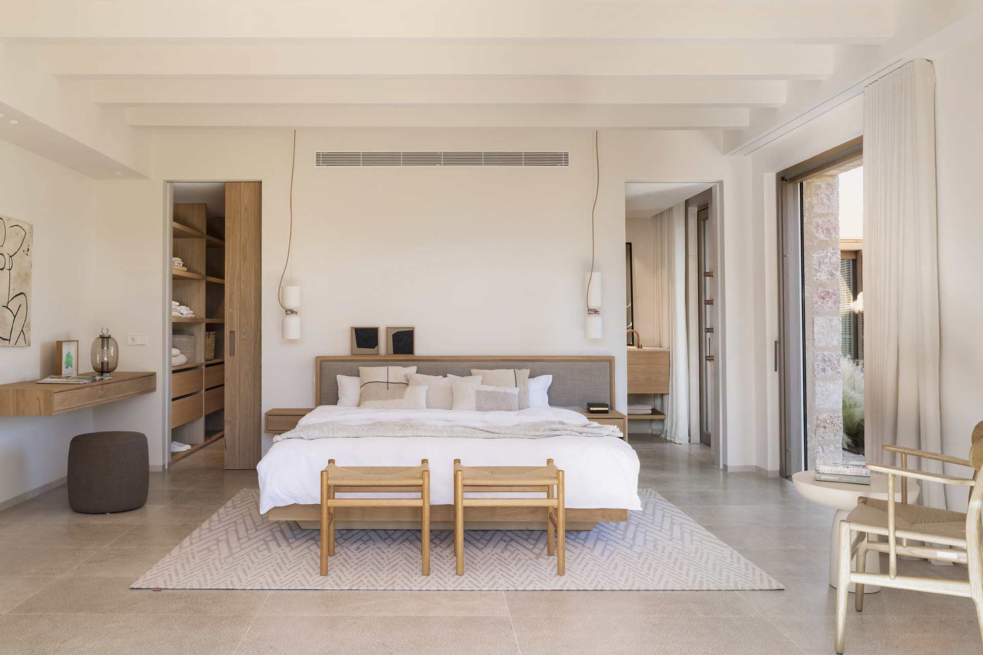 Dormitorio con cama de madera y cabecero tapizado, vestidor abierto y suelo de piedra clara
