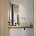 Lavabo de piedra sobre encimera de madera, con espejo alto, grifo de cobre y paredes de microcemento
