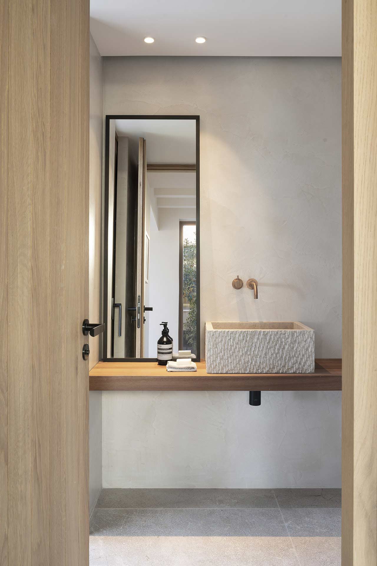 Lavabo de piedra sobre encimera de madera, con espejo alto, grifo de cobre y paredes de microcemento