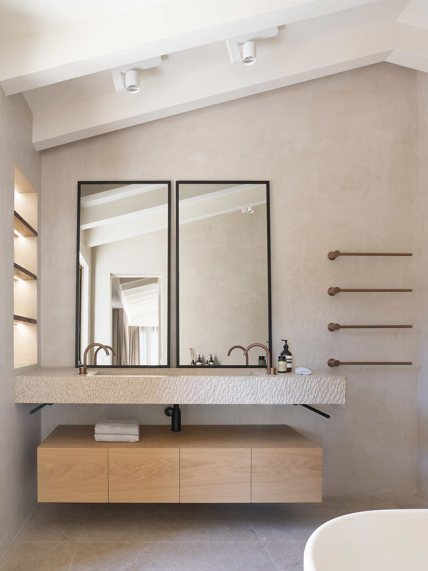 Baño con doble lavabo de piedra, espejos de marco negro, toalleros de cobre y mueble de madera