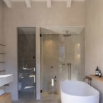 Baño con bañera exenta blanca, ducha acristalada revestida de piedra y grifería de cobre