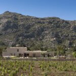 Villa de piedra a lo lejos, al pie de una gran ladera rocosa, tras hileras de viñedo