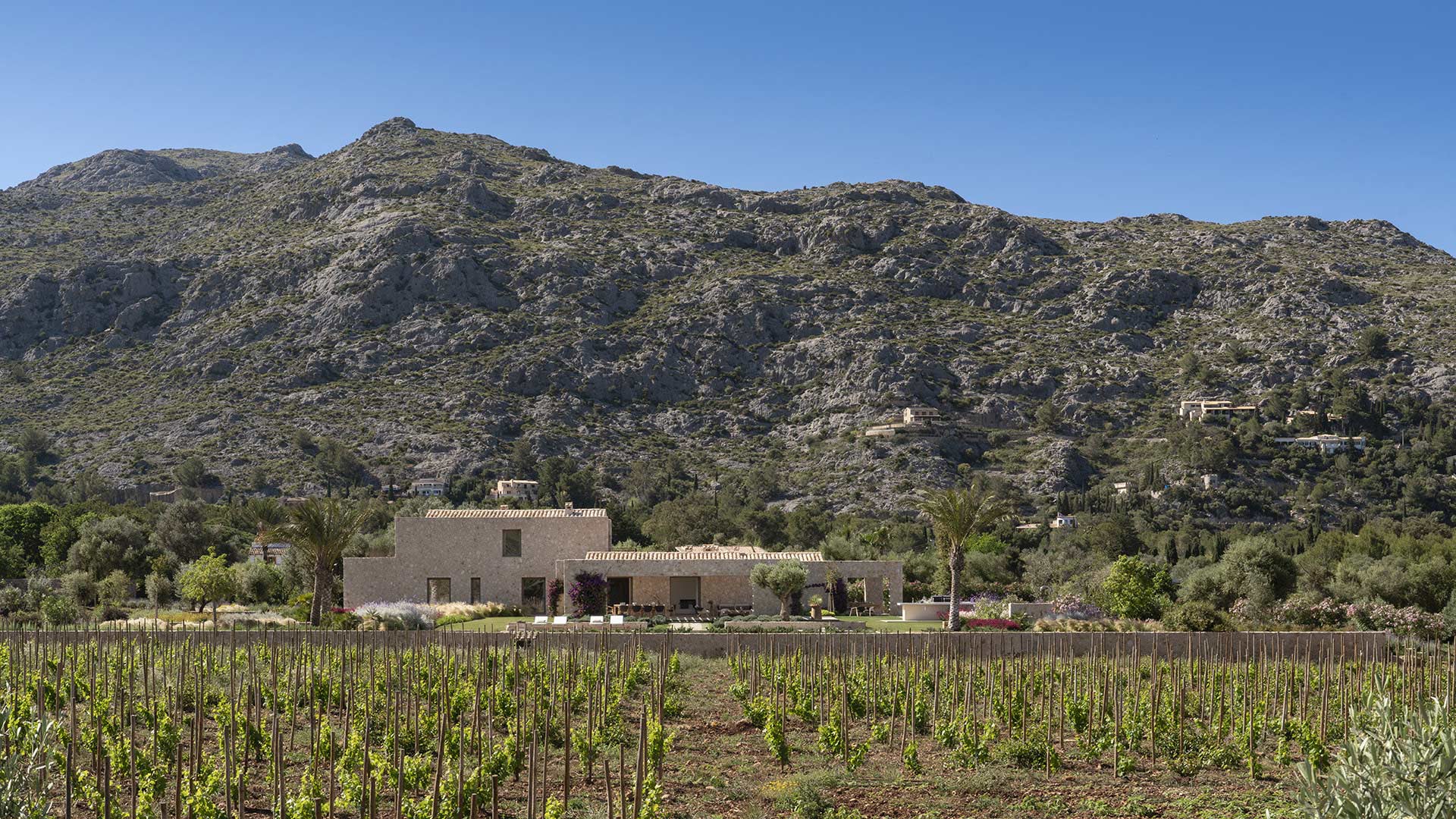 Villa de piedra a lo lejos, al pie de una gran ladera rocosa, tras hileras de viñedo