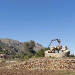 Pozo de piedra con mecanismo de madera sobre una base circular, con olivo y casas al fondo