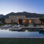Villa de piedra iluminada al anochecer, con piscina, tumbonas, reflejos en el agua y montañas