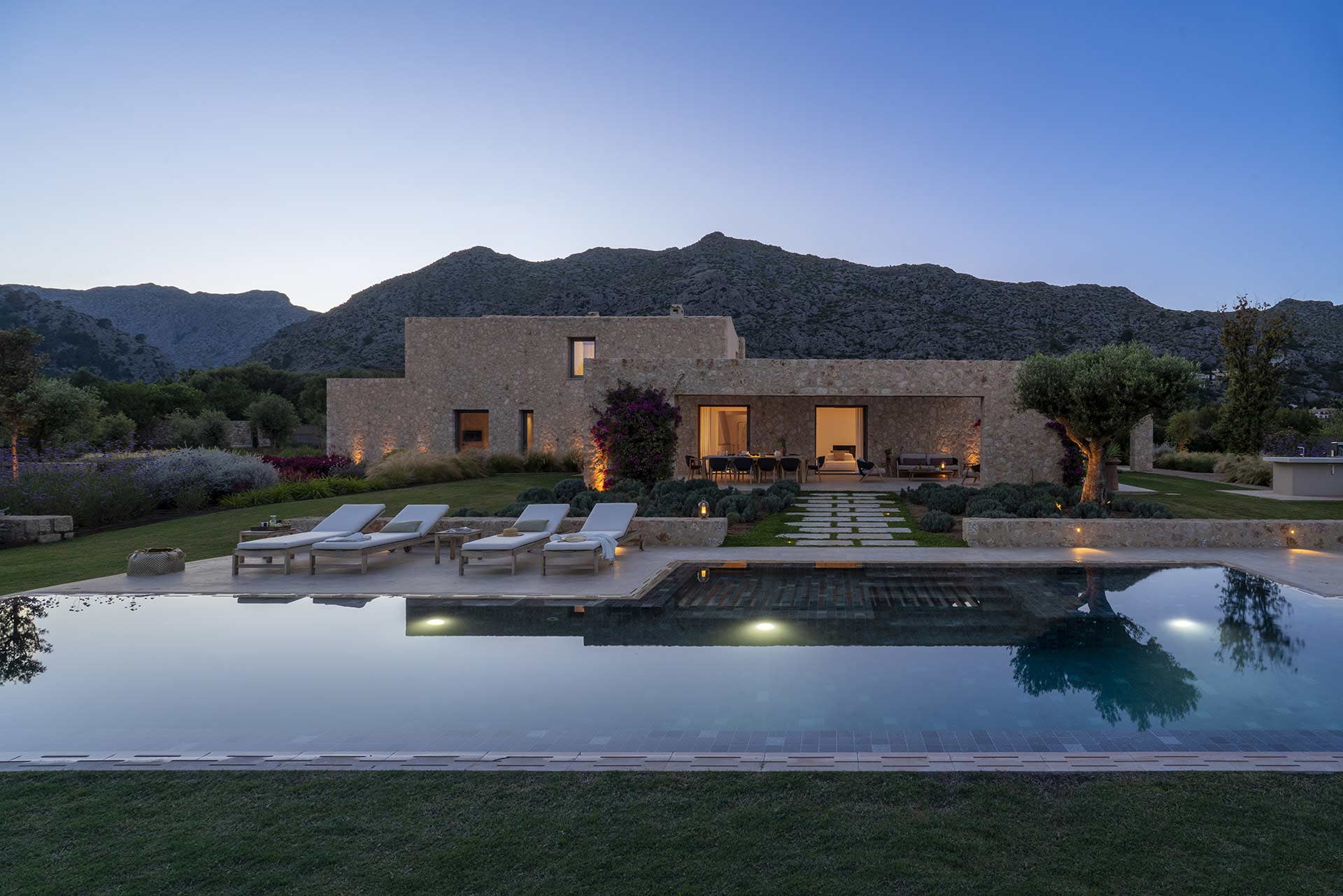 Villa de piedra iluminada al anochecer, con piscina, tumbonas, reflejos en el agua y montañas