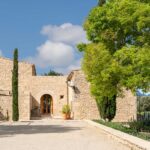 Entrada de piedra con puerta de madera en arco, ciprés alto y patio de gravilla soleado