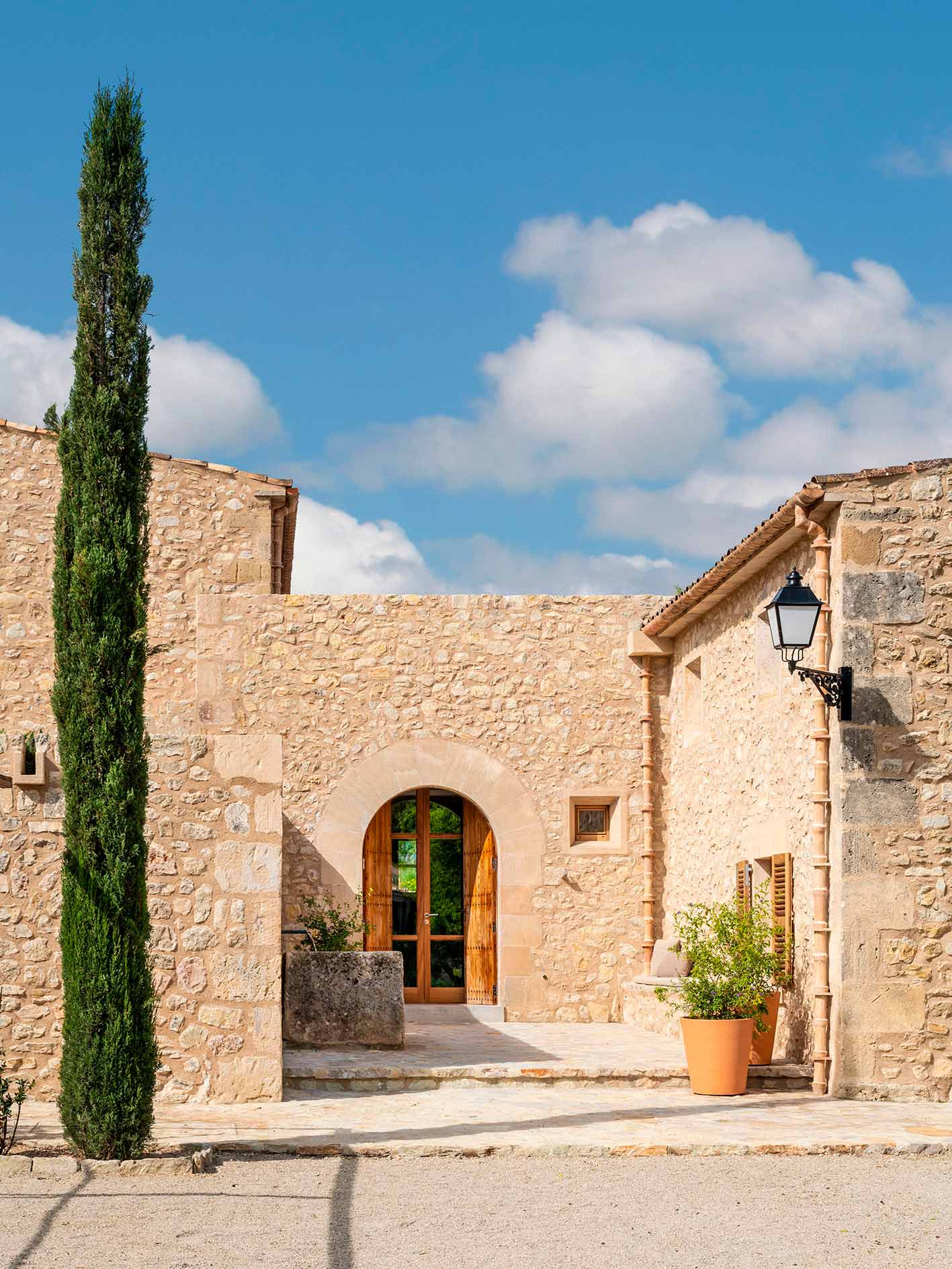 Detalle de la puerta en arco de madera entre muros de piedra, con ciprés, maceta y farol