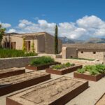 Huerto con bancales de acero corten, riego por goteo y casa de piedra con montañas al fondo