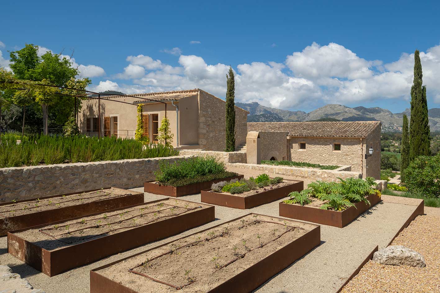Huerto con bancales de acero corten, riego por goteo y casa de piedra con montañas al fondo