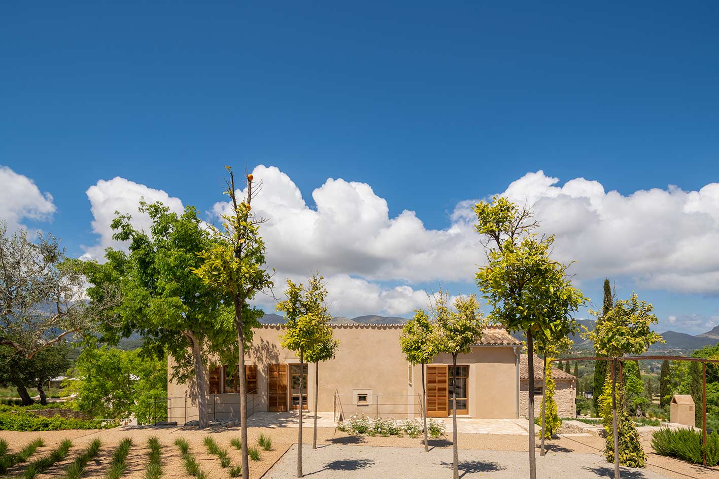 Casa de fachada color arena con contraventanas de madera, arbolitos jóvenes y montañas al fondo