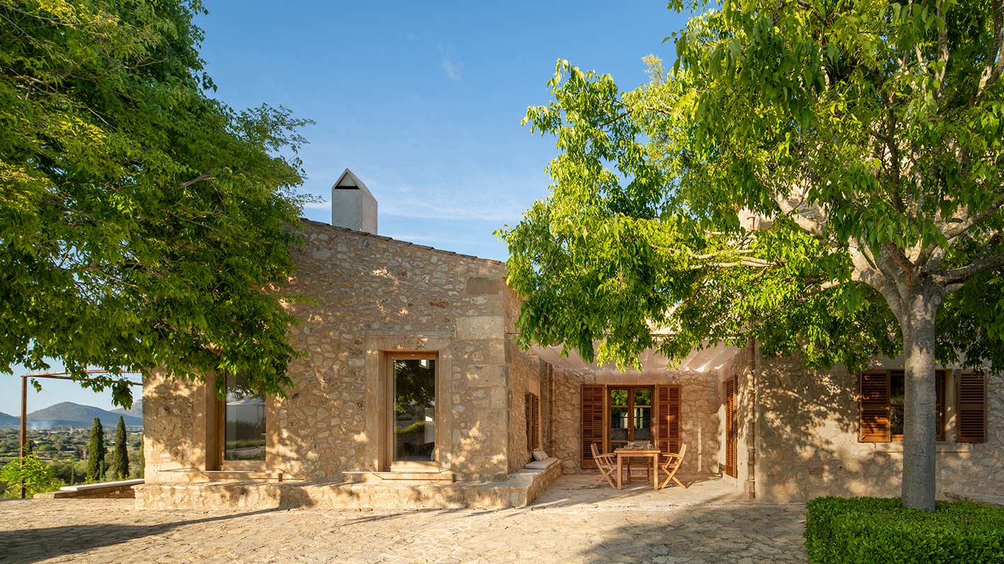 Porche de piedra con mesa y sillas plegables de madera, chimenea y grandes árboles de sombra