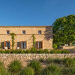 Fachada de piedra al sol con pérgola metálica, contraventanas de madera y muro de piedra seca