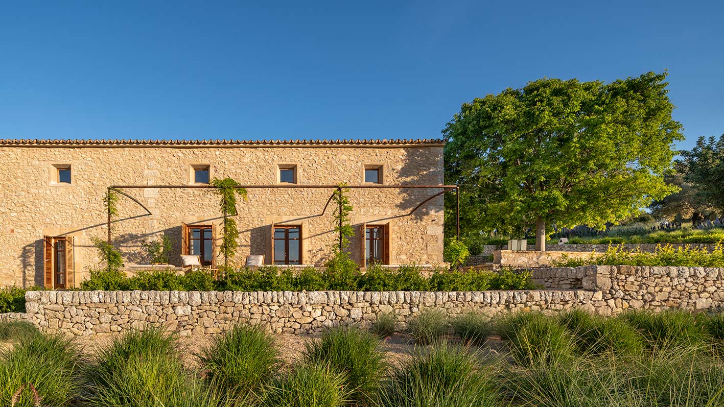 Fachada de piedra al sol con pérgola metálica, contraventanas de madera y muro de piedra seca