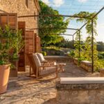 Terraza de piedra con mecedora de madera, cojines claros, pérgola metálica con enredadera y contraventanas