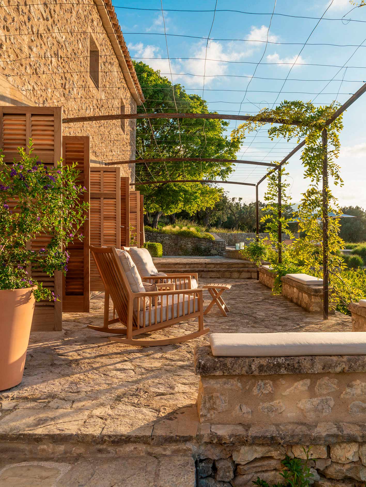 Terraza de piedra con mecedora de madera, cojines claros, pérgola metálica con enredadera y contraventanas