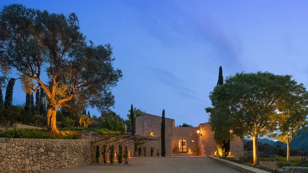 Casa rural Can Murta al anochecer, muros de piedra, olivo iluminado y montañas al fondo
