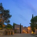 Casa rural Can Murta al anochecer, muros de piedra, olivo iluminado y montañas al fondo