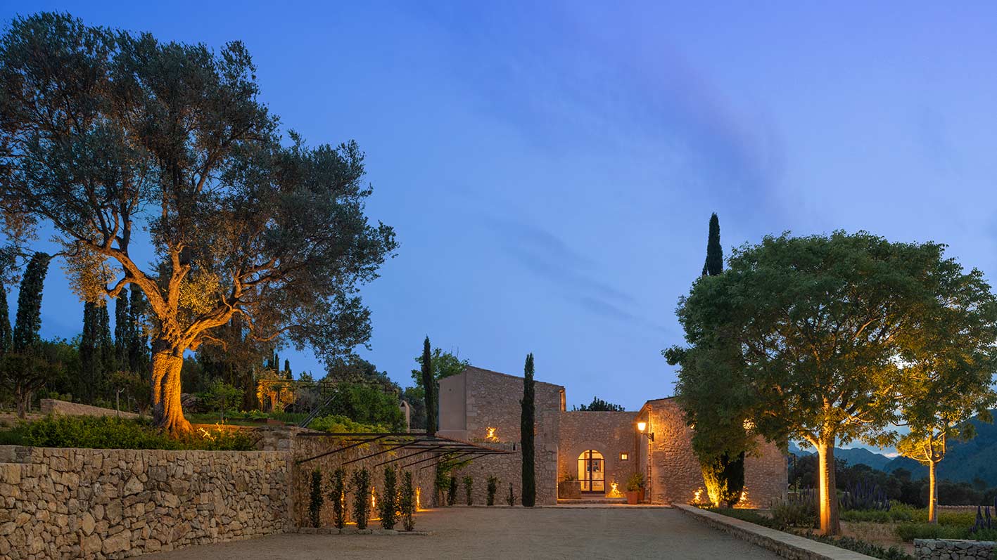 Casa rural Can Murta al anochecer, muros de piedra, olivo iluminado y montañas al fondo