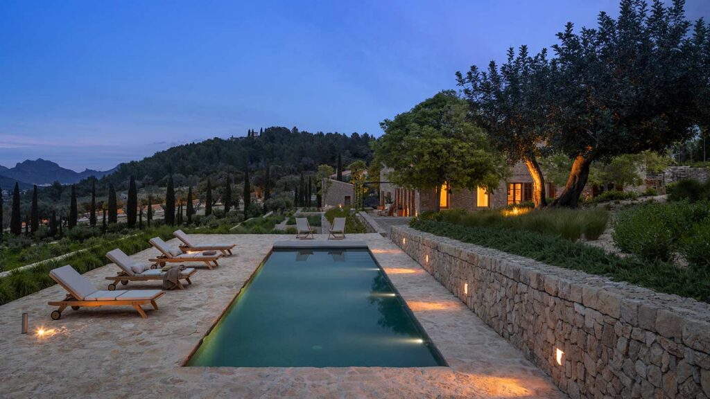 Piscina alargada iluminada al anochecer, tumbonas de madera, muro de piedra y montañas al fondo