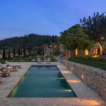 Piscina alargada iluminada al anochecer, tumbonas de madera, muro de piedra y montañas al fondo
