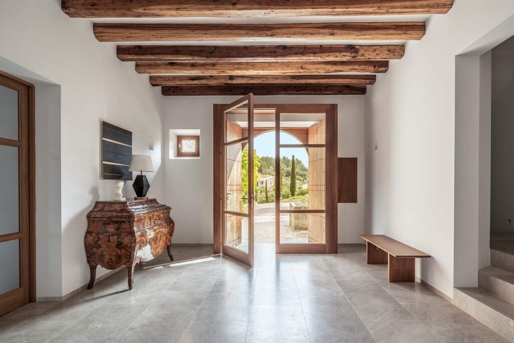 Recibidor con vigas de madera vista, suelo de piedra gris, cómoda antigua y puerta acristalada abierta al exterior