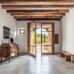 Recibidor con vigas de madera vista, suelo de piedra gris, cómoda antigua y puerta acristalada abierta al exterior