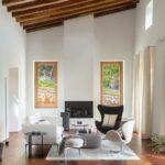 Salón de techo alto con vigas de madera, suelo de espiga, chimenea, butacas y cortinas claras