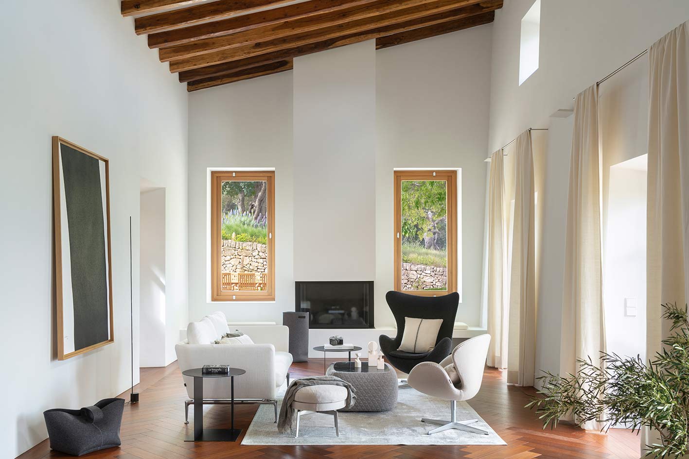 Salón de techo alto con vigas de madera, suelo de espiga, chimenea, butacas y cortinas claras