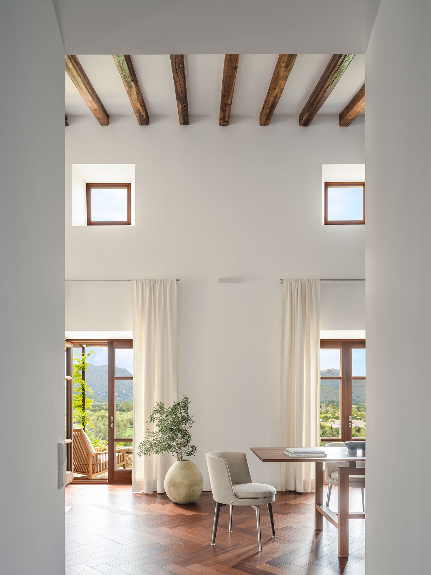 Sala de doble altura con ventanas altas, cortinas claras y puertas acristaladas con vistas a la montaña